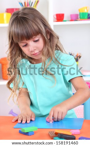 Little girl molds from plasticine sitting at table in room on shelves background