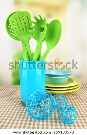 Plastic kitchen utensils in stand with clean dishes on tablecloth on bright background