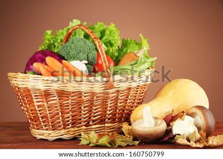 Different vegetables in basket with yellow leaves on table on brown background