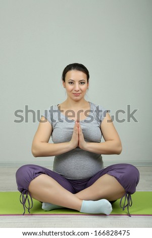 Young pregnant woman sitting and practicing yoga on mat on wall background
