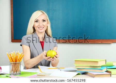 School teacher with apple sitting at table on blackboard background