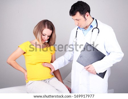 Young pregnant woman sitting on hospital bed with doctor on gray background