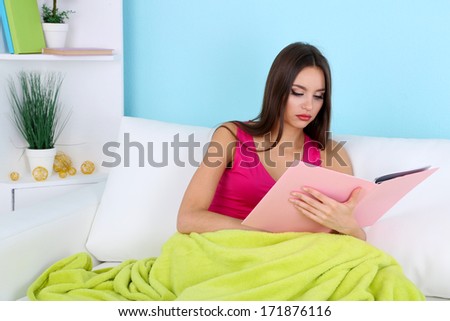 Beautiful young woman reading book on sofa on blue background