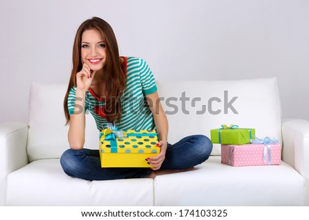 Beautiful young woman sitting on sofa with gift box on gray background