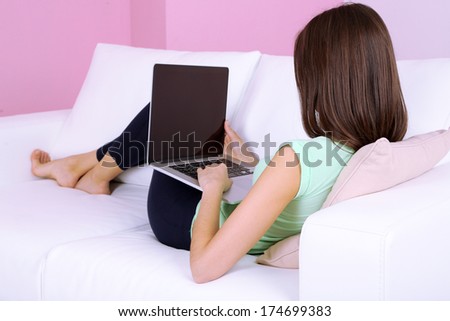 Young woman sitting on sofa with laptop on pink background