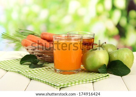 Glasses of juice, apples and carrots on white wooden table, on green background