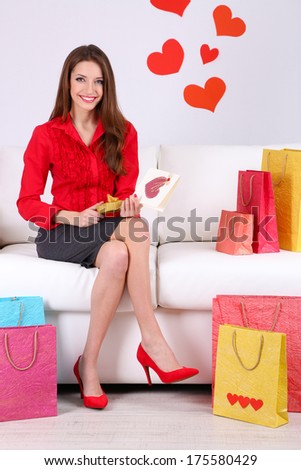 Beautiful young woman sitting on sofa with shopping bags and gift box on gray background