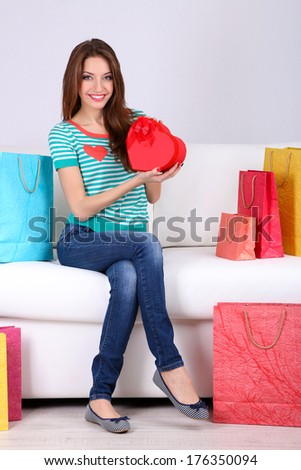 Beautiful young woman sitting on sofa with shopping bags and gift box on gray background