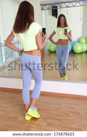 Pretty sporty girl photographing herself in mirror at phone in fitness room