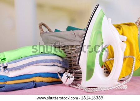 Iron and pile of colorful clothes and basket on table on bright background