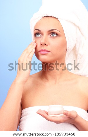 Beautiful young woman after shower with a towel on her head and a jar of cream in hand on blue background close-up