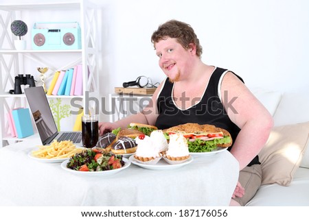 Fat man has a big lunch and playing games on laptop, on home interior background
