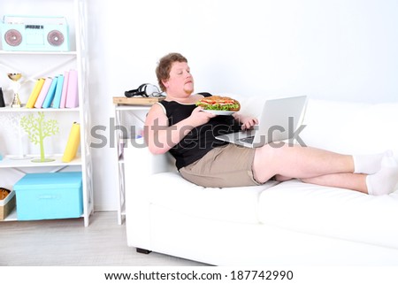 Fat man sitting with laptop on sofa and eating tasty sandwich on home interior background