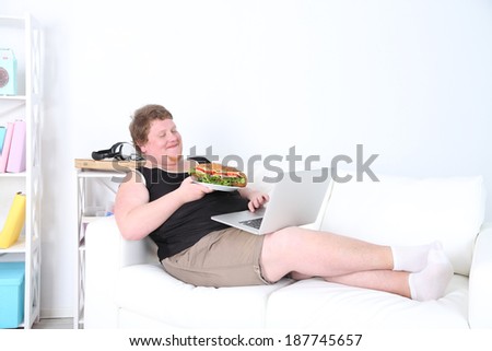 Fat man sitting with laptop on sofa and eating tasty sandwich on home interior background