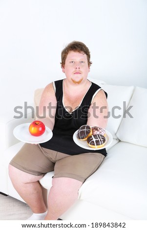 Fat man holding plated with apple and donuts on home interior background.  Choosing between good healthy food and bad unhealthy food