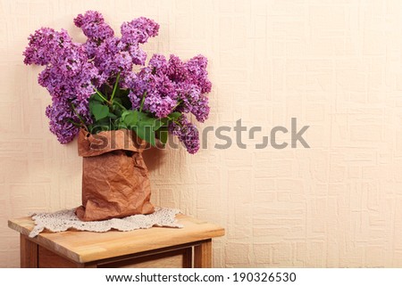 Beautiful lilac flowers in paper bag on wooden table near wall