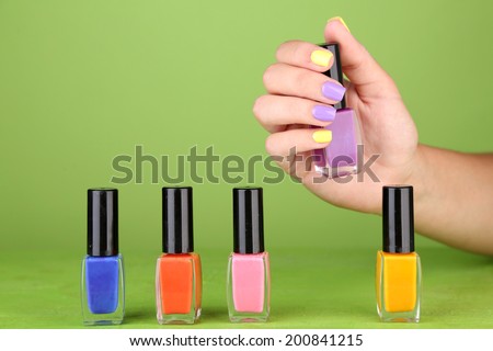 Female hand with stylish colorful nails holding bottle with nail polish, on green background
