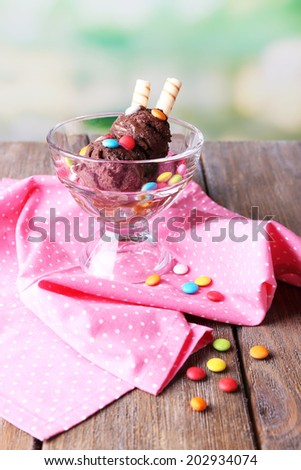 Chocolate ice cream with multicolor candies and wafer rolls in glass bowl, on color wooden table, on bright background