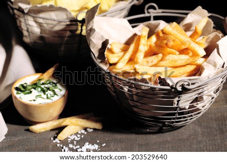 Tasty french fries in metal basket and potato chips on wooden table with dark light