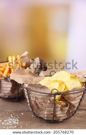 Tasty potato chips and french fries in metal baskets on wooden table, on light background