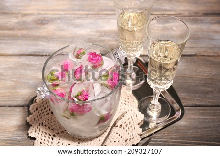 Ice cubes with rose flowers in glass bucket and two glasses with champagne on wooden table background