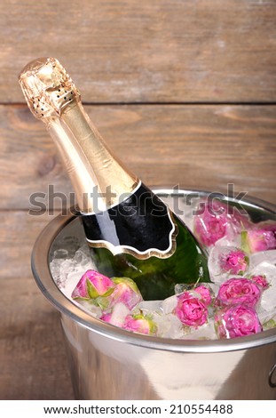 Frozen rose flowers in ice cubes and champagne bottle in bucket, on wooden background