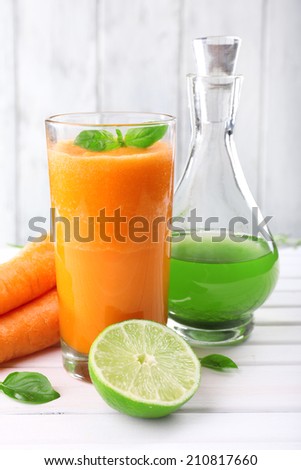 Glass of fresh carrot juice, decanter of lime juice, lime,  carrot, tuft of grass on the table in front of wooden wall
