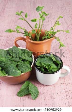 Brown round bowl and white metal mug of fresh mint leaves and cup of mint branches on pink wooden background