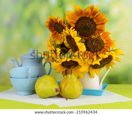 Beautiful sunflowers in pitcher with cups and pears on table on bright background