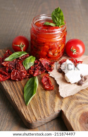 Sun dried tomatoes in glass jar, basil leaves and feta cheese on cutting board, on wooden background