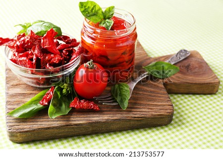 Sun dried tomatoes in glass jar, basil leaves on cutting board, on table background