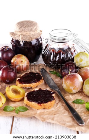 Plum jam, slices of bread with plum jam and fresh plums in glass dish on piece of paper on wooden table on light background
