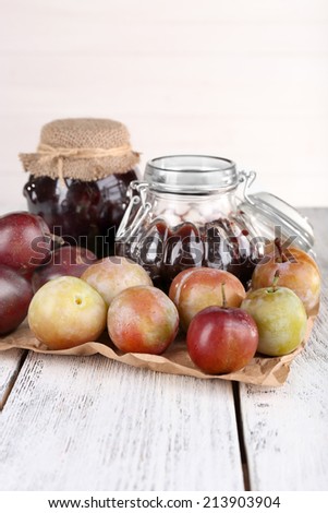 Plum jam and fresh plums in glass dish on piece of paper on wooden table on light background