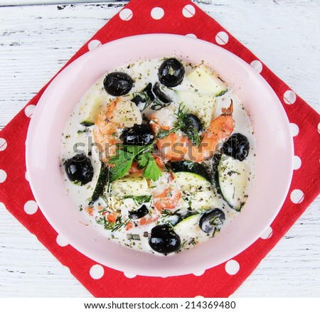 Fresh prawns with olives, avocado and parsley in white sauce in a pink round plate on a napkin on wooden background