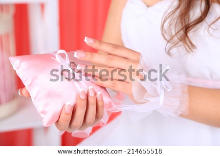 Bride in white dress and gloves holding decorative pillow with wedding rings, close-up, on bright background