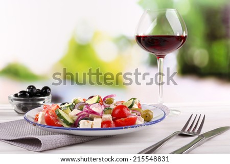 Greek salad served in plate with glass of wine on napkin on wooden table on natural background