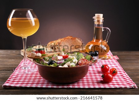 Bowl of Greek salad served with olive oil and glass of wine on napkin on wooden table on dark background