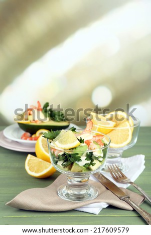 Tasty salads with shrimps and avocado in glass bowl and on plate, on wooden table, on bright background