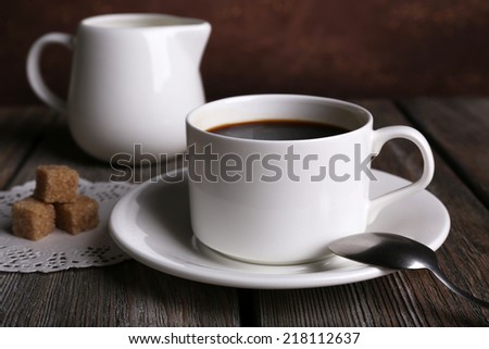 Cup of coffee with cream in milk jug and sugar cubes on wooden table on dark background