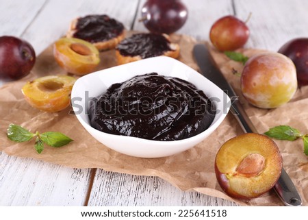 Plum jam, slices of bread with plum jam and fresh plums in glass dish on piece of paper on wooden table on light background