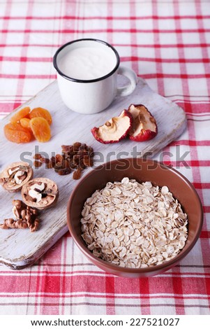 Bowl of oatmeal, mug of yogurt, marmalade, chocolate, raisins, dried apricots and walnuts on wooden cutting board on checkered fabric background