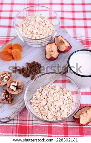 Oatmeal in bowls, mug of yogurt, marmalade, chocolate, raisins, dried apricots and walnuts on wooden cutting board on checkered fabric background