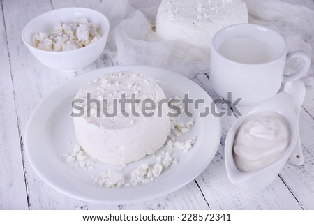 Cottage cheese on plate on gauze and cottage cheese in bowl, cup of milk and cream in saucer boat on wooden background