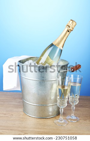 Bottle of champagne in metal ice bucket and two glasses on wooden table on color background