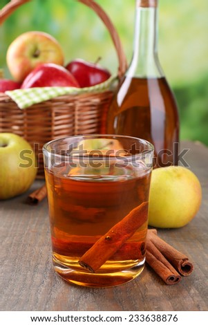 Apple cider in glass and bottle, with cinnamon sticks and fresh apples on wooden table, on bright background