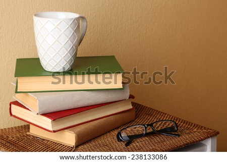 Pile of books with cup and glasses on wicker surface and light wall background