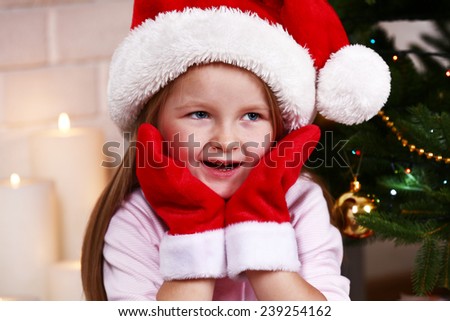 Little girl in Santa hat and mittens sitting near fir tree on fireplace with candles background
