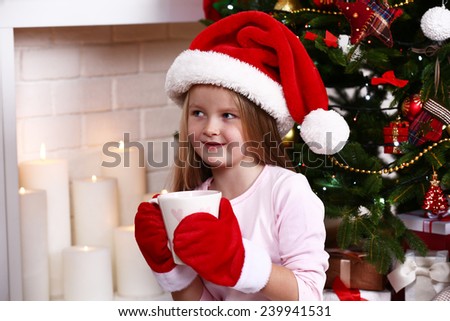 Little girl in Santa hat and mittens taking cup sitting near fir tree on fireplace with candles background