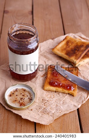 Toast bread spread with jam on piece of paper with knife near jar on wooden table background
