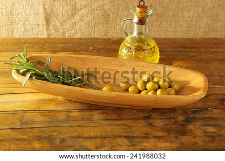 Green olives and branch in oblong bowl with oil can on rustic wooden table, on burlap background